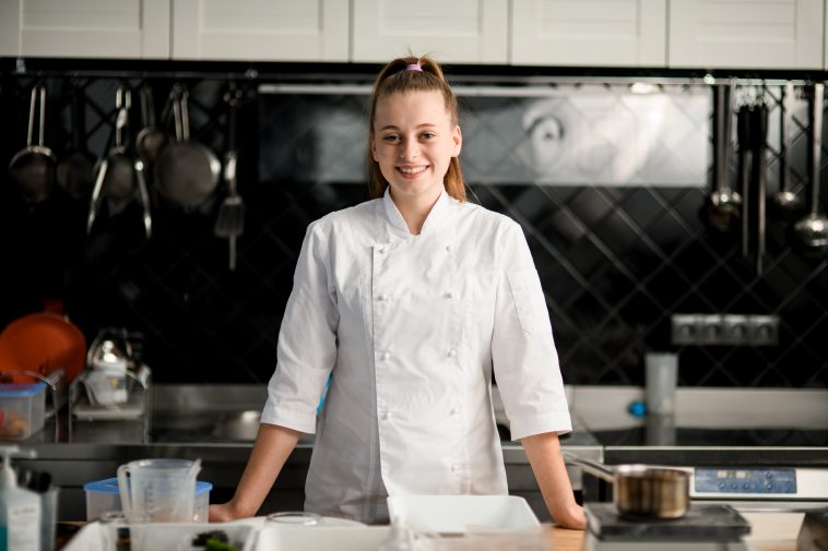 view on handsome young smiling woman chef wearing in white suit at kitchen. Blurred background