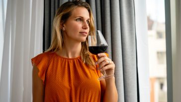 Beautiful young woman with glass of red wine standing, relaxing near window