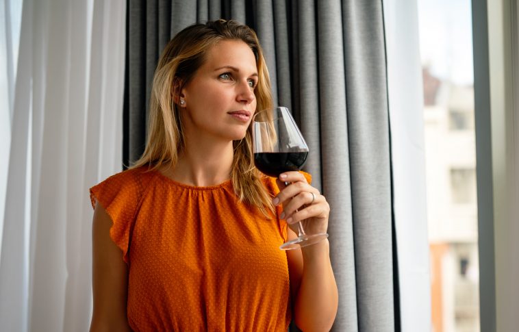 Beautiful young woman with glass of red wine standing, relaxing near window