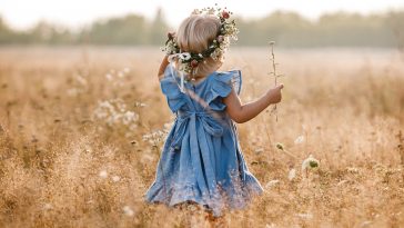 little girl is wearing a flower wreath on her head in a field on summer sunny day. baby in a blue dress.Portrait of adorable little child outdoors. happy holiday childhood. back view.