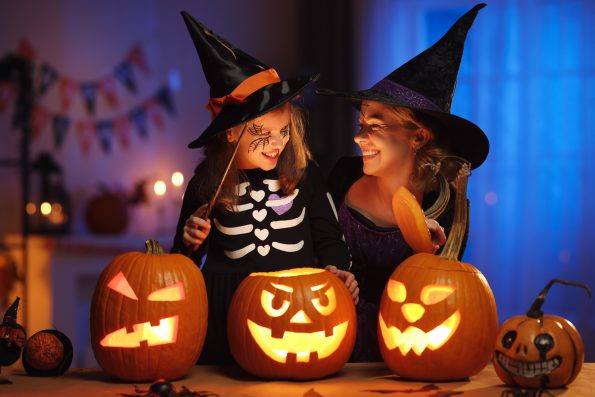 Happy family mother and little girl daughter in witch costumes celebrating Halloween holiday together at home, mom and child with magic wand playing in dark house room decorated for All hallows eve