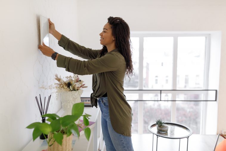 Modern Home Interior And Domestic Decor. Smiling young black woman hanging painting, putting photo picture frame on the wall. Casual lady taking care of coziness in her new stylish apartment, profile