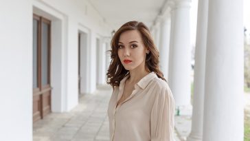 pretty caucasian young woman in beige blouse, seductive red lipstick and wavy hair standing on veranda of cafe. white concrete pillars in background