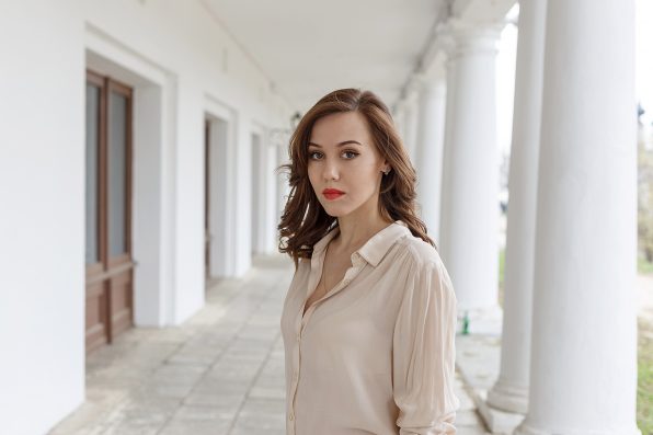 pretty caucasian young woman in beige blouse, seductive red lipstick and wavy hair standing on veranda of cafe. white concrete pillars in background