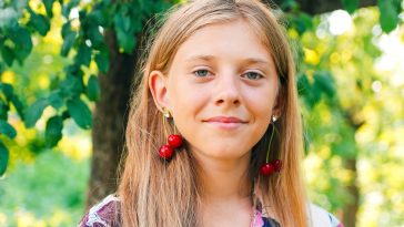 Defocus beautiful blond teen girl with sweet cherry on ear. Beautiful smiling teenage girl closeup, against green of summer park. Child earrings. Summer portrait. Out of focus.