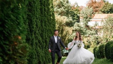 Front view of cheerful loving brides walking in the green garden, beautiful bride holding bouquets of flowers wearing wedding dress. Married couple looking at each other.