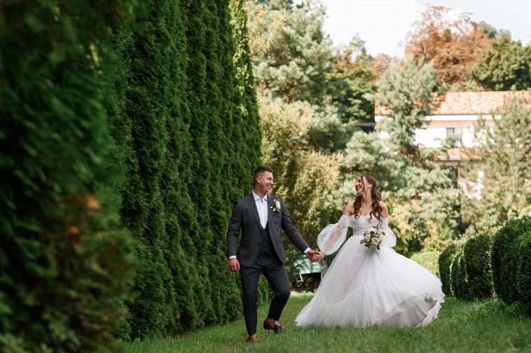 Front view of cheerful loving brides walking in the green garden, beautiful bride holding bouquets of flowers wearing wedding dress. Married couple looking at each other.