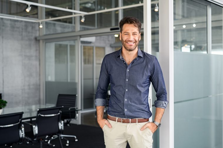 Portrait of mature successful man with hands in pocket standing outside office conference room. Handsome businessman standing in office while looking at camera. Young satisfied man in formalwear standing in boardroom with copy space.