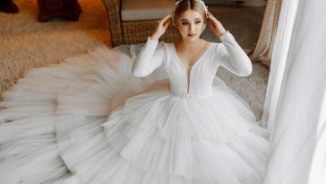 Portrait of a beautiful bride in a white wedding dress with a long train in a hotel room. The bride is sitting on the floor against a large window