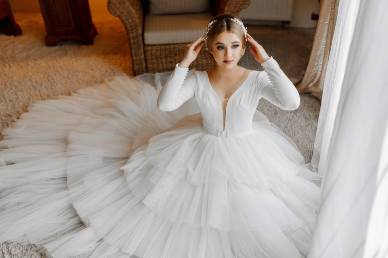 Portrait of a beautiful bride in a white wedding dress with a long train in a hotel room. The bride is sitting on the floor against a large window