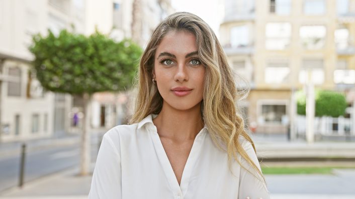 Cool, relaxed young blonde woman seriously looking at camera, standing casually in sunny urban street, attractive lifestyle portrait, outdoors with city background