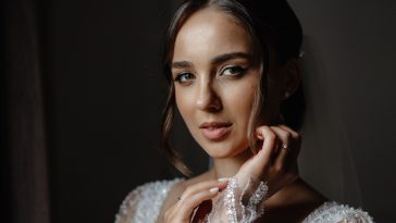 A young woman with long, dark hair and smokey eye makeup is wearing a white wedding dress with lace sleeves and a sweetheart neckline. She is looking at the camera with a soft smile.