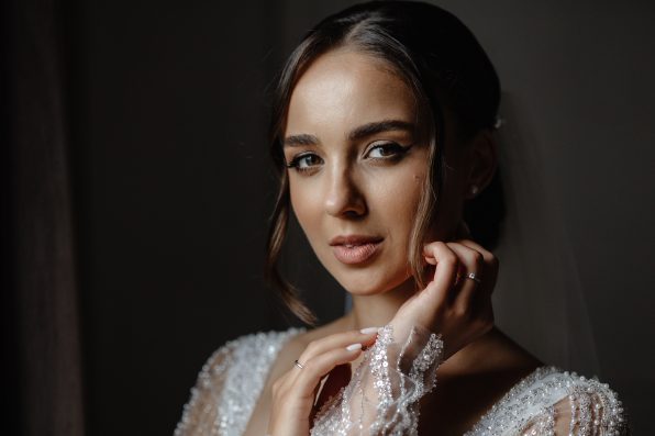 A young woman with long, dark hair and smokey eye makeup is wearing a white wedding dress with lace sleeves and a sweetheart neckline. She is looking at the camera with a soft smile.