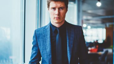 Half length portrait of serious expert economist in elegant suit keeping hands in pockets, stylish confident businessman in formal wear looking at camera while standing in modern company office