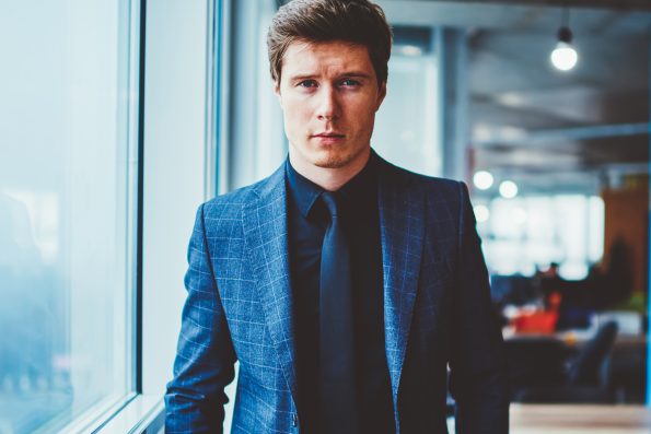 Half length portrait of serious expert economist in elegant suit keeping hands in pockets, stylish confident businessman in formal wear looking at camera while standing in modern company office