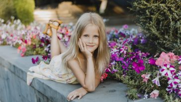 portrait of a cute girl 5-6 years old with a petunia flower, sitting over flowering bushes in the garden outdoors. Spring season. Stylish little child against the backdrop of nature. Spring. Childhood.