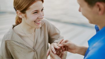 Smiling woman receiving engagement ring from man on rooftop during sunset, while wearing casual attire and displaying joyful expressions