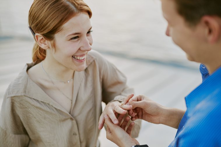 Smiling woman receiving engagement ring from man on rooftop during sunset, while wearing casual attire and displaying joyful expressions