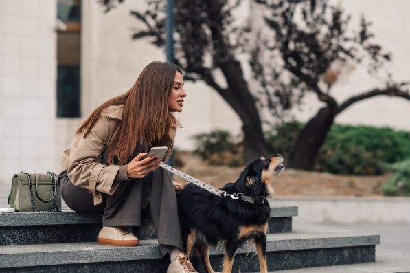 Businesswoman working remotely outside, her dog barking in the city. Despite distractions, she stays focused on her phone, balancing work and pet ownership