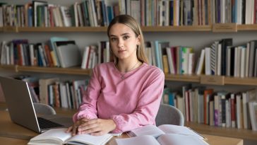 Serious beautiful teenage Caucasian student girl preparing for college exam in high school public library, sitting at table with heap of books and laptop, looking at camera for portrait