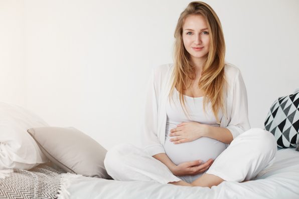 Healthy pregnancy gestation. Portrait of a young pregnant smiling woman sitting in her bedroom resting in the morning holding her tummy waiting for her childbirth.