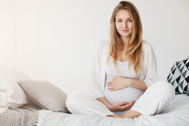 Healthy pregnancy gestation. Portrait of a young pregnant smiling woman sitting in her bedroom resting in the morning holding her tummy waiting for her childbirth.