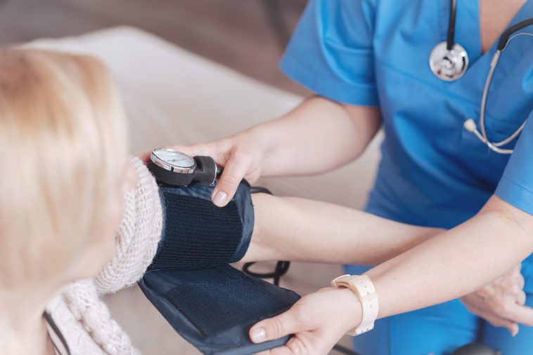 Treatment and prevention. Scaled up look on a mature nurse sitting next to a blonde lady and fixing a sphygmomanometer on her arm before a regular pressure check up.