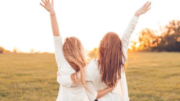 Two pretty girls raised their hands on a field at sunset.