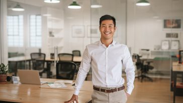 Smiling young Asian businessman standing next to a table while working alone in a large modern office