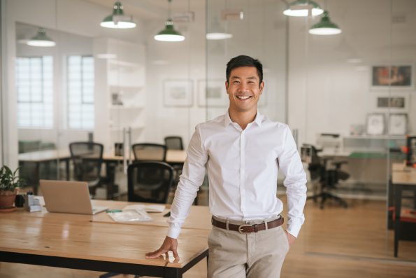 Smiling young Asian businessman standing next to a table while working alone in a large modern office