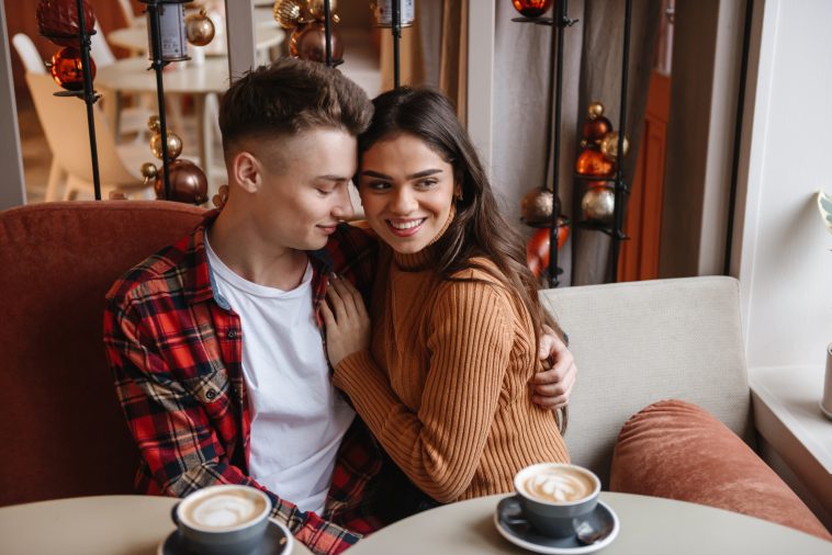 Picture of a cute young happy loving couple sitting in cafe indoors hugging.