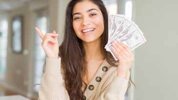 Young woman holding 50 dollars bank notes very happy pointing with hand and finger to the side