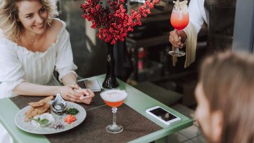 Charming blond lady looking at young man with smile while bartender holding glass of cocktail. Couple sitting at the table with food, bouquet of reed berries and smartphones