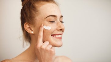 Close up of a young female with red hair and freckles applying white cream on her face laughing with closed eyes isolated on white background.