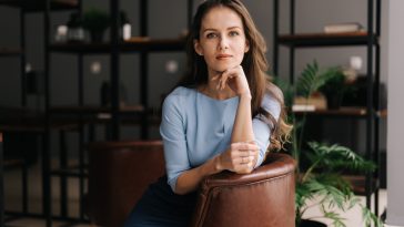 Beautiful young businesswoman wearing stylish light blue dress sitting at the chair in office with modern interior, looking at the camera. Portrait of charming business lady.