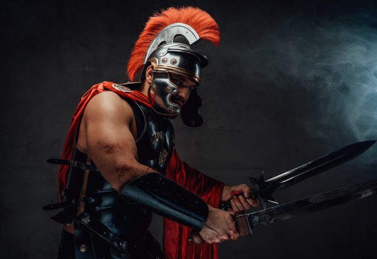 Armoured savage and brutal roman warrior in red cloak with helmet attacks holding swords in smokey background.