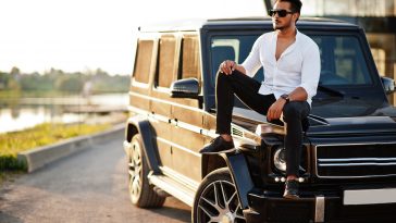 Solid asian man in white shirt and sunglasses posed near black mafia suv car.