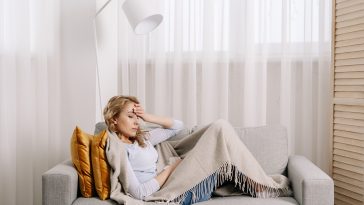 Concept of health problem. Sick and unhappy woman covered with plaid, touching forehead, suffering from strong headache or chronic migraine. Female sit on couch at home feeling pain in head