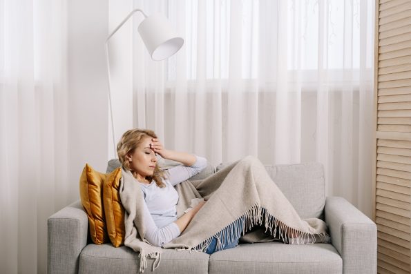 Concept of health problem. Sick and unhappy woman covered with plaid, touching forehead, suffering from strong headache or chronic migraine. Female sit on couch at home feeling pain in head