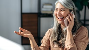 Satisfied successful middle aged gray-haired lady, business woman,  have phone talk with coworker or friend, gesturing with hand, looks away and happily smile