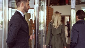 Helpful doorman wearing uniform welcoming people inside the hotel while holding the entrance door open