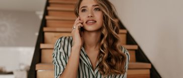 Charming curly woman in green and white silk striped shirt sits on wooden stairs, talks on cellphone and smiles at home.