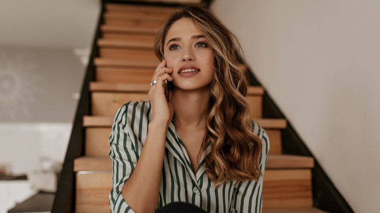 Charming curly woman in green and white silk striped shirt sits on wooden stairs, talks on cellphone and smiles at home.