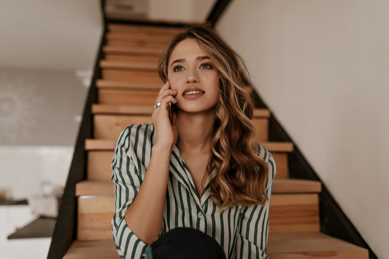 Charming curly woman in green and white silk striped shirt sits on wooden stairs, talks on cellphone and smiles at home.