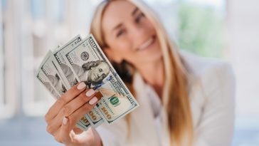 Dollar banknotes in hands of blurry cheerful beautiful blonde businesswoman offering money. Business salary and profit concept. Swedish female young woman holding money happy to get benefits.