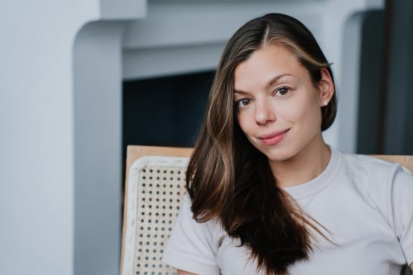 Portrait of adorable brunette girl in white t-shirt sitting at home in chair against fireplace. Confident caucasian young adult woman smiles looks at camera. Satisfied successful lady relaxing indoors