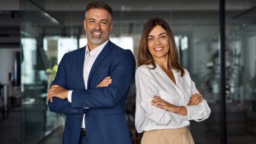 Portrait of smiling mature Latin or Indian business man and European business woman standing arms crossed in office. Two diverse colleagues, group team of confident professional business people.