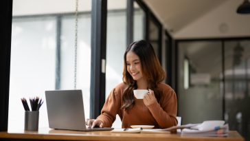 A young woman takes enjoy a hot beverage, with a smile, at her sunny office desk.