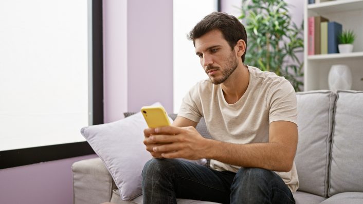 A focused young hispanic man with a beard using a smartphone while sitting in a modern living room.