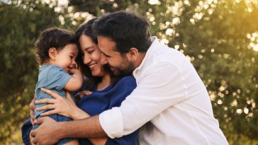 A joyful family moment with a father, mother in a blue dress, and their one-year-old son in blue clothes, hugging and smiling in a grassy natural setting with a tree in the background